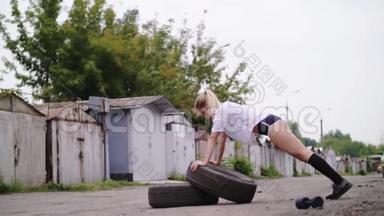 运动年轻的金发女人穿短裤，在轮胎、俯卧撑的帮助下进行各种力量练习。